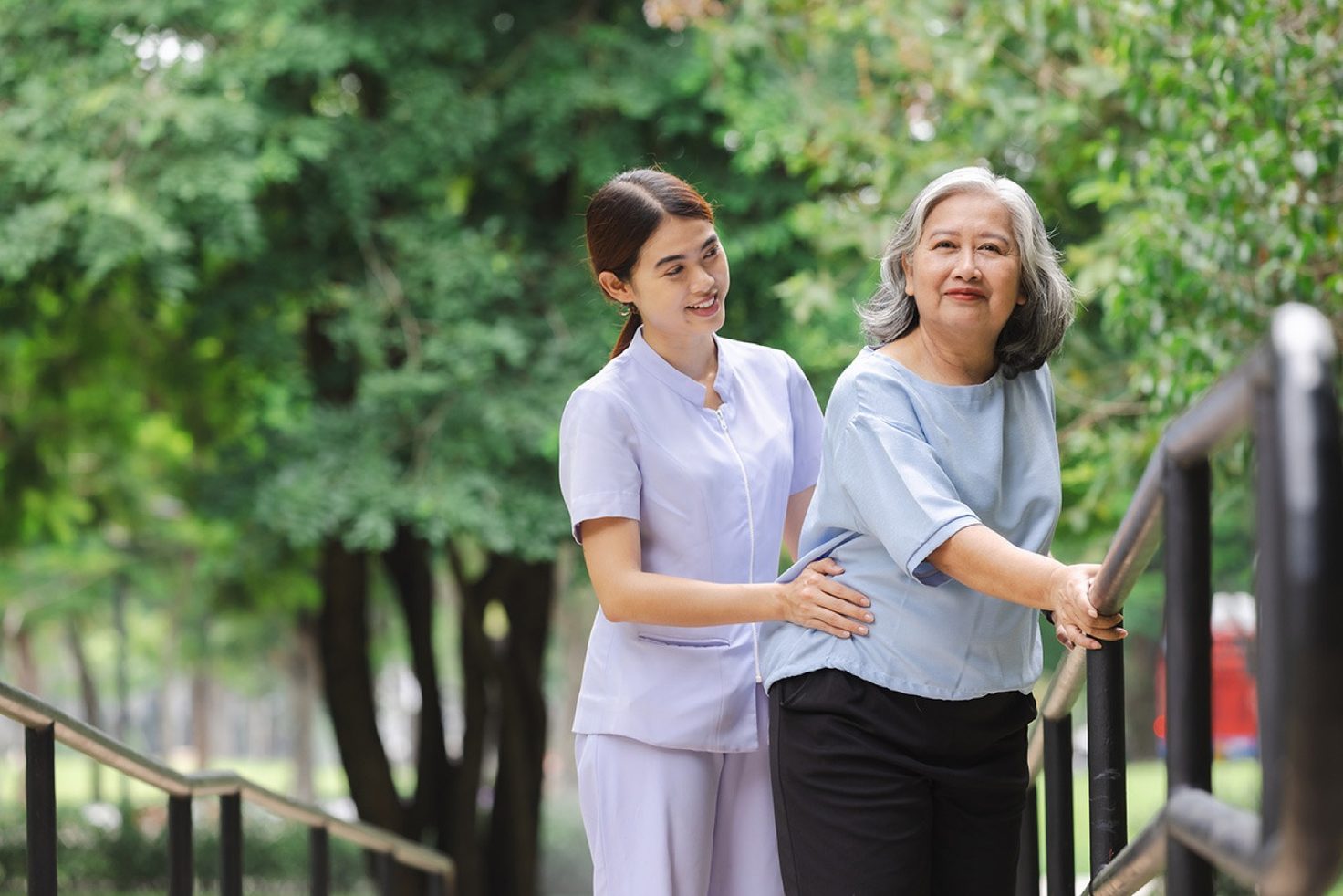 A caregiver steadies an older woman by the hip as she walks along an outdoor railing