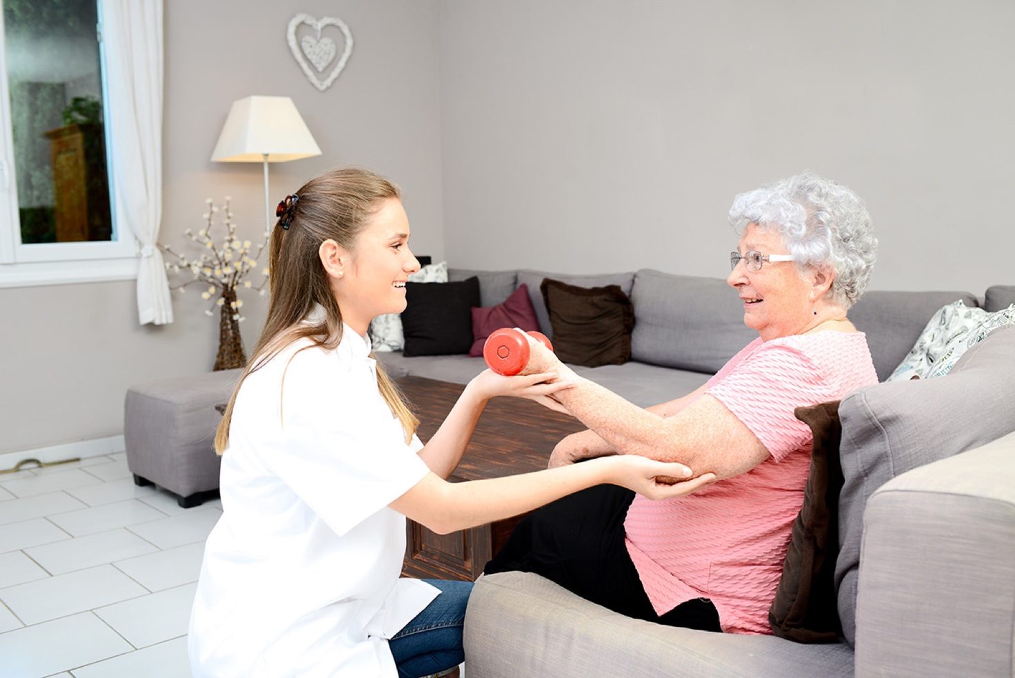 A clinician hands a red dumbbell to an older woman exercising on her couch at home