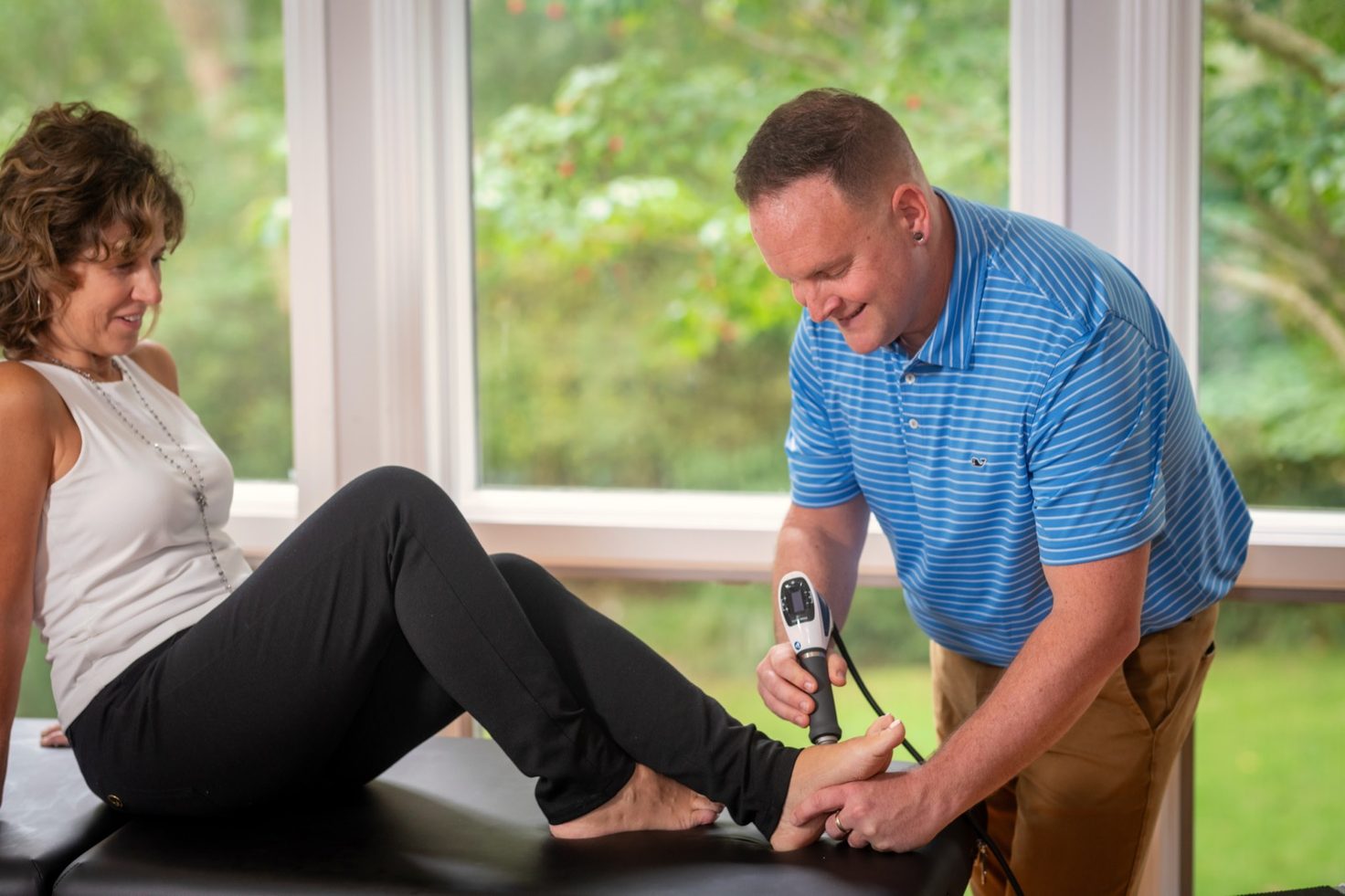 A physical therapist applies a handheld shockwave device to a patient's foot on a treatment table