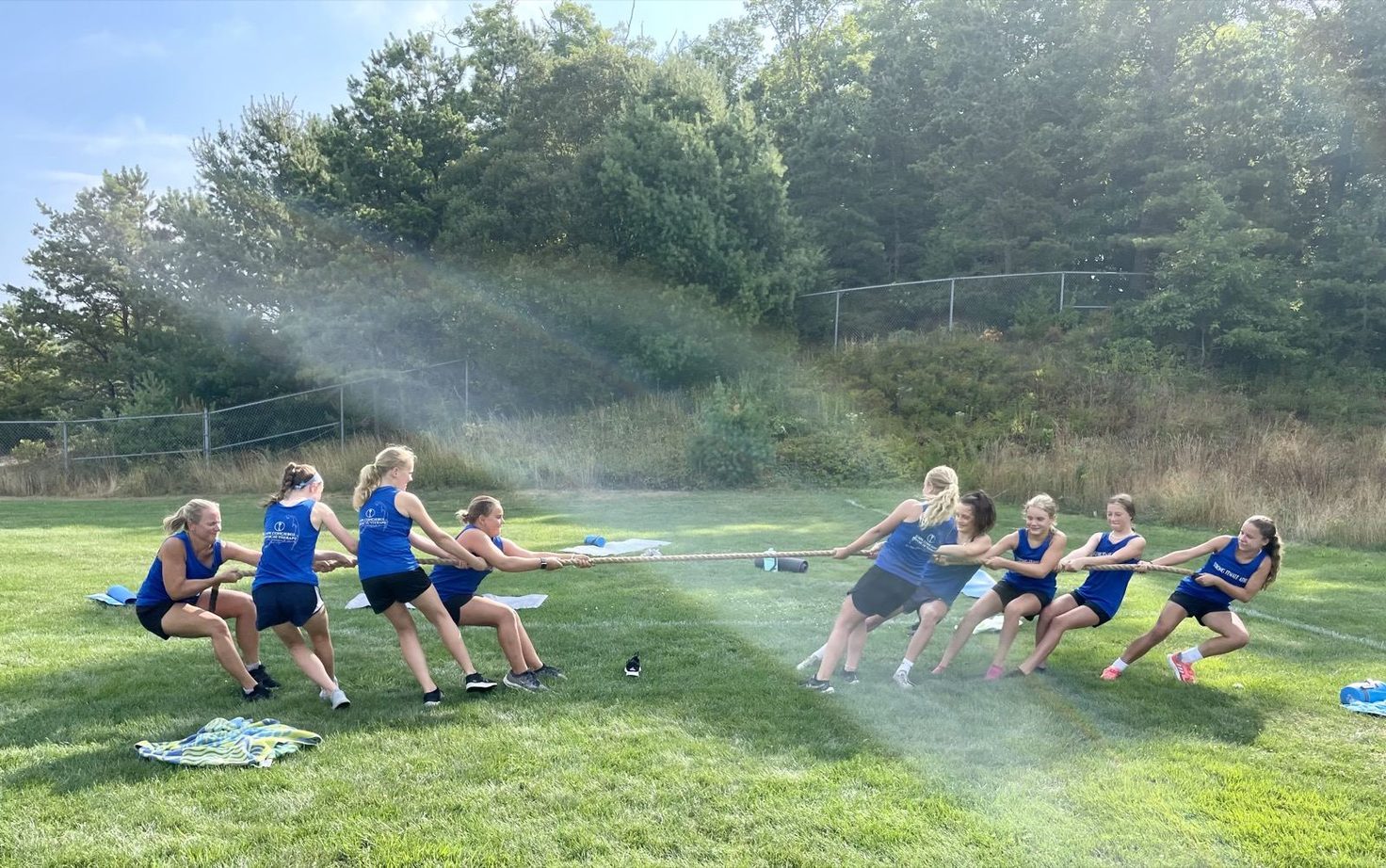 Two teams of girls and women in blue shirts play tug-of-war on a sunny grass field